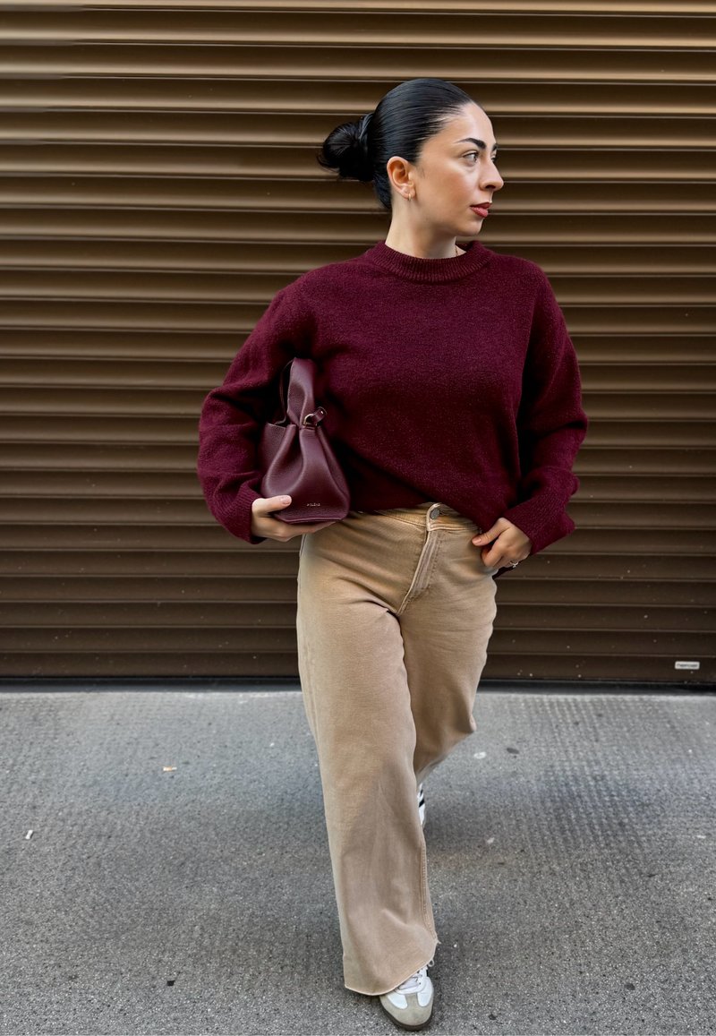 Pull en maille bordeaux avec un col montant, un pantalon beige à jambes larges et un petit sac à main bourgogne. Les baskets sont crèmes avec un accent gris.