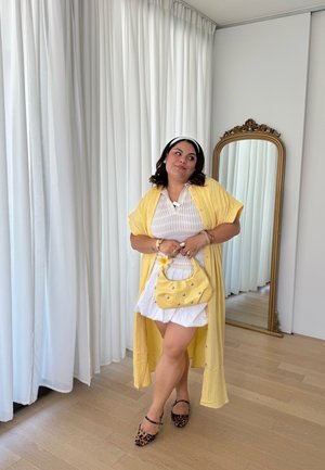 Kimono largo amarillo sobre una blusa blanca y pantalones cortos, llevando un bolso amarillo con tachuelas. Zapatos planos de estampado de leopardo, diadema. Espejo de cuerpo entero al fondo.