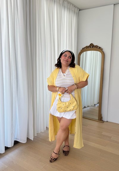 Kimono largo amarillo sobre una blusa blanca y pantalones cortos, llevando un bolso amarillo con tachuelas. Zapatos planos de estampado de leopardo, diadema. Espejo de cuerpo entero al fondo.