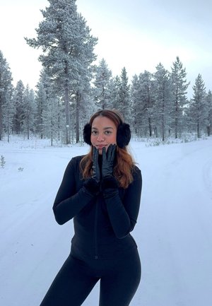 Joven mujer con atuendo de invierno negro, orejeras y guantes, de pie en una carretera nevada con pinos cubiertos de nieve en el fondo.