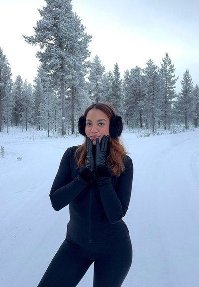Joven mujer con atuendo de invierno negro, orejeras y guantes, de pie en una carretera nevada con pinos cubiertos de nieve en el fondo.