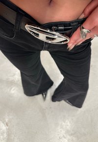 Pessoa vestindo calças pretas largas e um cinto preto com uma grande fivela prateada, ajustando o cinto na altura da cintura, usando um anel prateado na mão.