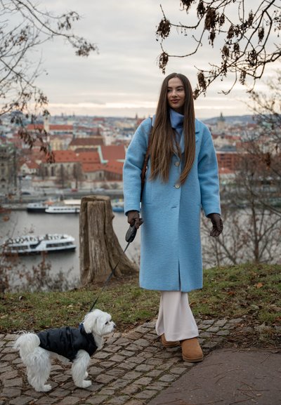 Abrigo de lana azul con corte recto y detalle de botones, combinado con pantalones crema y botas marrones. Un pequeño perro blanco con una chaqueta negra en la correa.