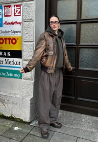 Barna bőr bomber dzseki, olívazöld kapucnis pulóver, laza szénszürke nadrág és barna cipők. A háttérben színes feliratok és texturált ablak látható.