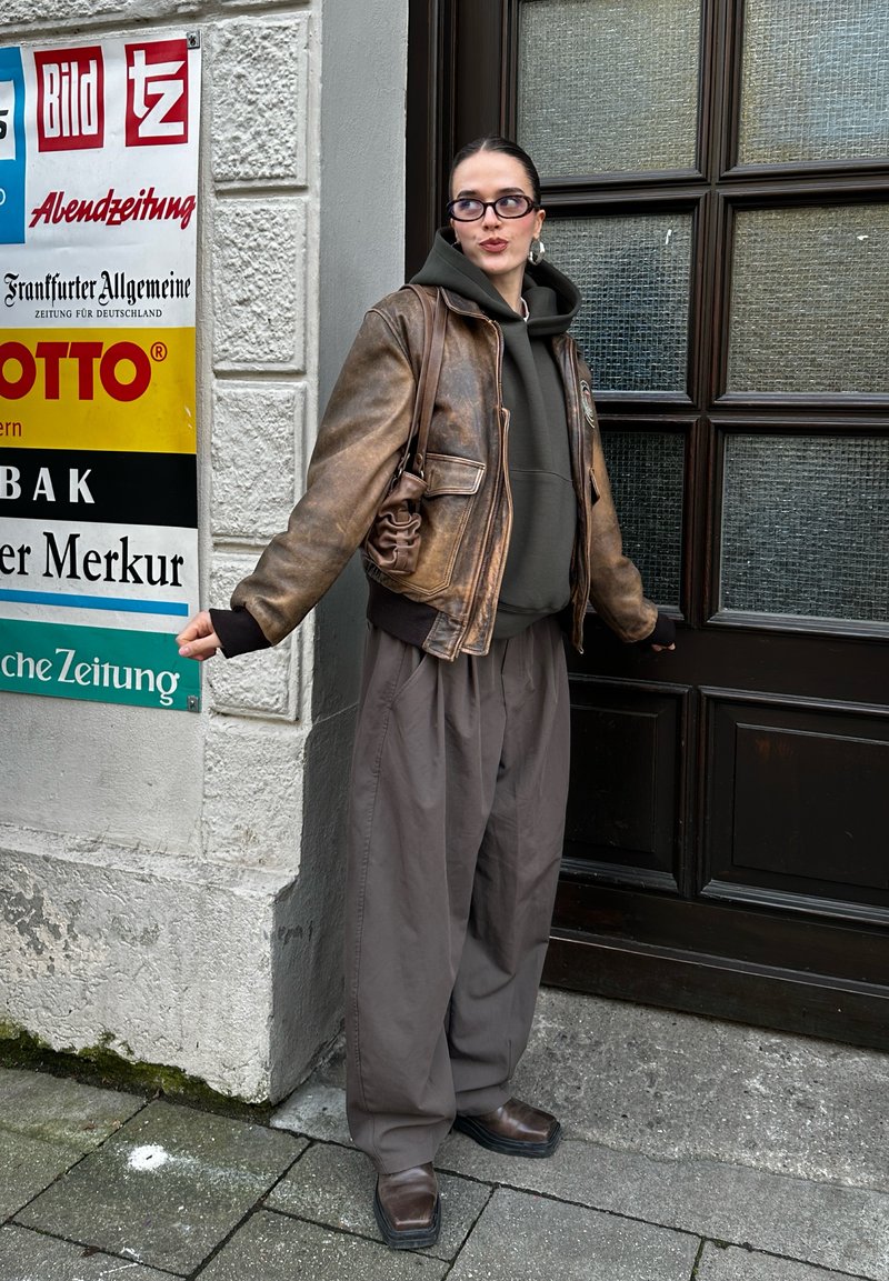 Giacca bomber in pelle marrone, felpa con cappuccio verde oliva, pantaloni larghi color carbone e scarpe marroni. Lo sfondo presenta insegne colorate e una finestra con texture.