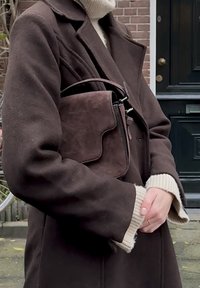 Geantă crossbody din piele întoarsă maro, cu clapetă curbată, accesorii metalice și o curea neagră, ținută aproape de o haină maro cu mâneci largi și manșete crem.