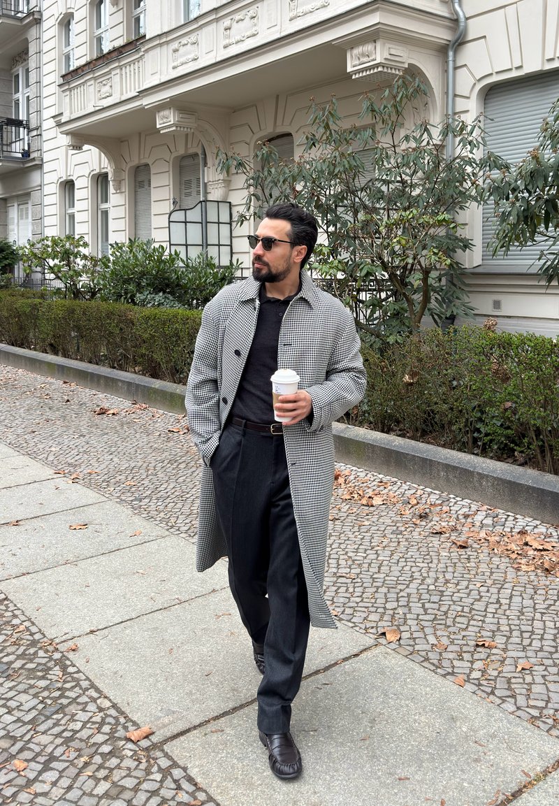 Homme portant un long manteau à carreaux, un pantalon noir et des lunettes de soleil marchant sur un trottoir en tenant un gobelet de café à emporter près de buissons feuillus et de bâtiments.