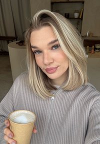 Jeune femme aux cheveux blonds portant un pull gris côtelé, tenant une tasse de café avec de la mousse, en intérieur avec des étagères et une décoration en arrière-plan.
