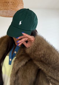 Άτομο με σκουρό πράσινο καπέλο, καφέ χνουδωτό παλτό και κίτρινο τοπ με μπλε γιακά, κρατώντας χέρι με μπλε νύχια κοντά στο πρόσωπο μέσα σε εσωτερικό χώρο.