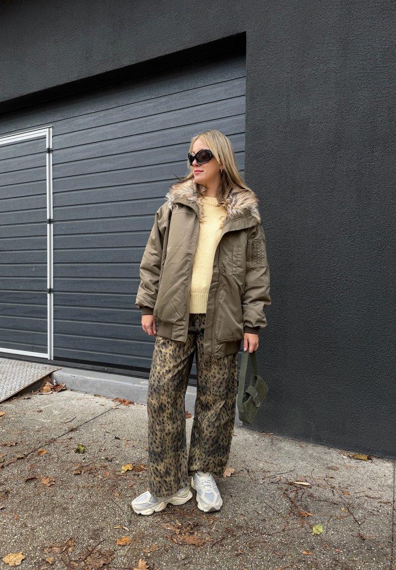 Femme portant une veste marron surdimensionnée bordée de fourrure, un pull jaune, un pantalon imprimé léopard, des baskets blanches, des lunettes de soleil et un sac à main vert, debout sur une surface pavée.