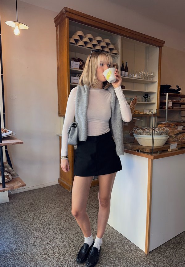 Femme avec une coupe au carré blonde boit dans une tasse blanche dans une boulangerie avec du pain et des bouteilles de vin sur les étagères derrière elle.
