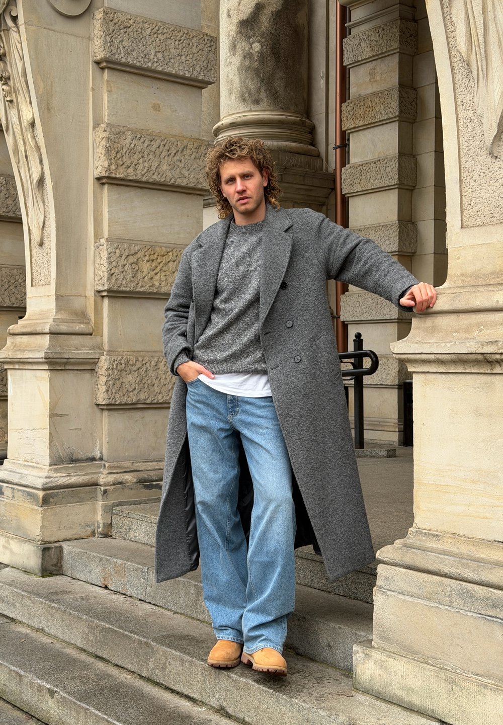 Long manteau en laine gris sur un pull gris, associé à un jean bleu clair et des chaussures beige ; debout sur des marches en pierre près d'une architecture ornée.