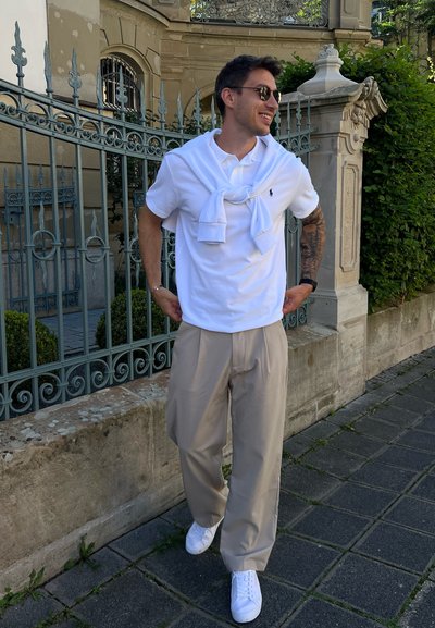 Camisa polo blanca combinada con un suéter blanco atado al cuello, pantalones beige y zapatillas blancas, de pie frente a una valla decorativa.