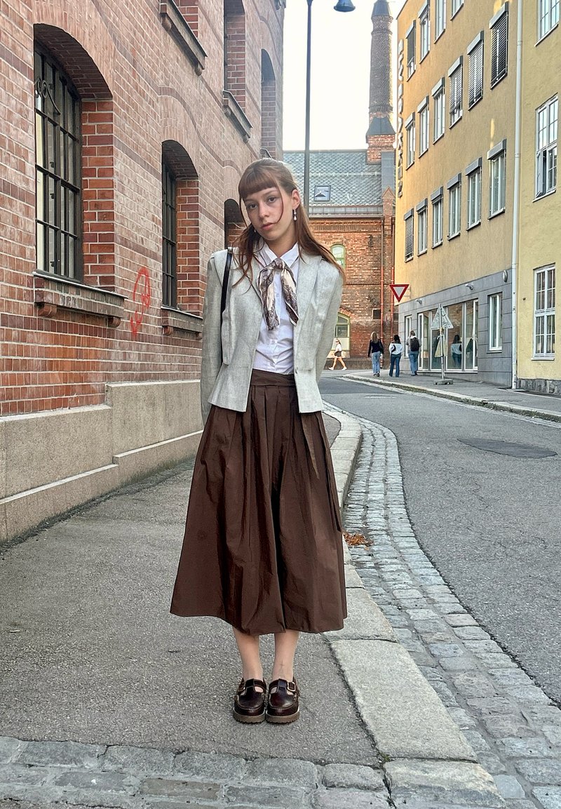 Jeune femme debout sur un trottoir en ville portant un chemisier blanc, une veste grise, une jupe marron et des chaussures foncées, avec des bâtiments en arrière-plan.