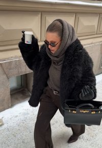 Kobieta w czarnym futrzanym płaszczu i szaliku trzyma filiżankę kawy oraz czarną torebkę, ostrożnie idąc po zaśnieżonej ulicy obok beżowego budynku.