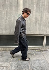Chaqueta de cuero marrón con cuello y bolsillos frontales, combinada con pantalones negros holgados y botines grises, caminando sobre una superficie pavimentada.
