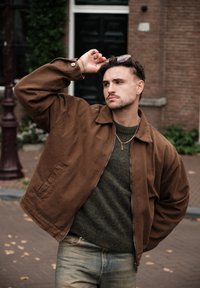 Jachetă bomber din bumbac maro cu fermoar, purtată peste un pulover verde închis. Prezintă o croială relaxată și un finisaj neted, texturat.
