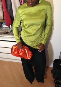Blusa de manga longa, brilhante, verde limão, combinada com calças de ganga escuras e largas, e uma clutch metálica laranja enrugada. Saltos pretos em bico visíveis.