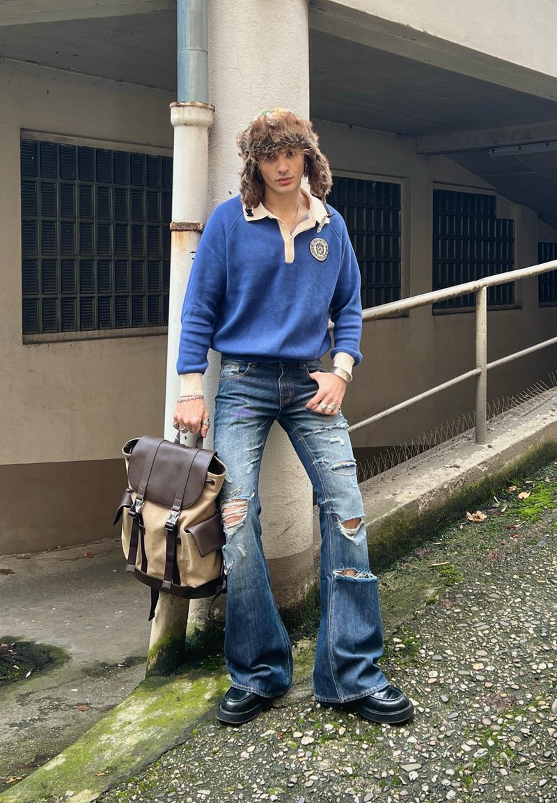 Blue fleece pullover with collar, emblem on the chest, light denim flared jeans with rips, brown and beige rucksack, black shoes, fur hat.