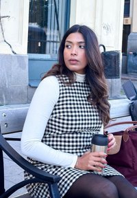 Uma mulher está sentada num banco, vestindo um vestido xadrez preto e branco por cima de uma blusa de manga longa branca, segurando um copo de viagem prateado.