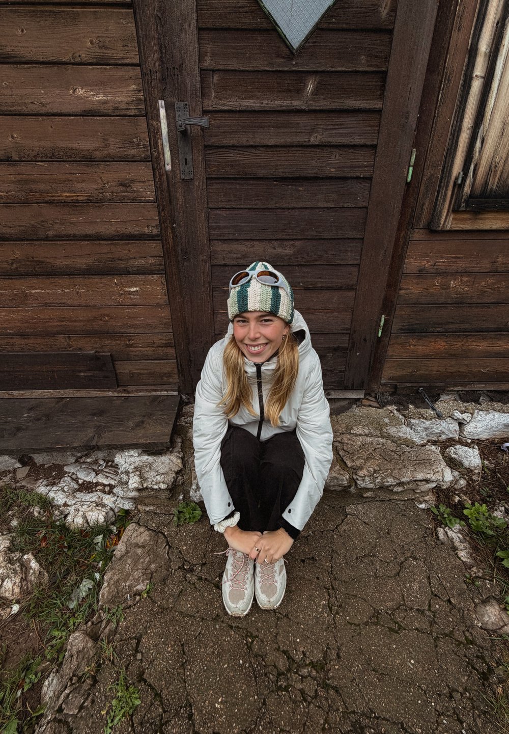 Hvid dunjakke, sorte bukser og grå vandresko med pink detaljer. Står foran en trædør og har en stribet strikhue på.