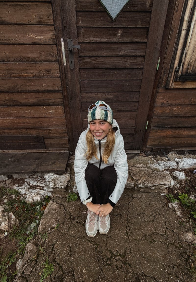 Doudoune blanche, pantalon noir et chaussures de randonnée grises avec des accents roses. Debout devant une porte en bois, portant un bonnet en laine rayé.