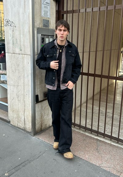 Joven con chaqueta negra, camisa a cuadros, pantalones negros y zapatos color canela, de pie cerca de una entrada con reja junto a un muro de concreto con un cartel.