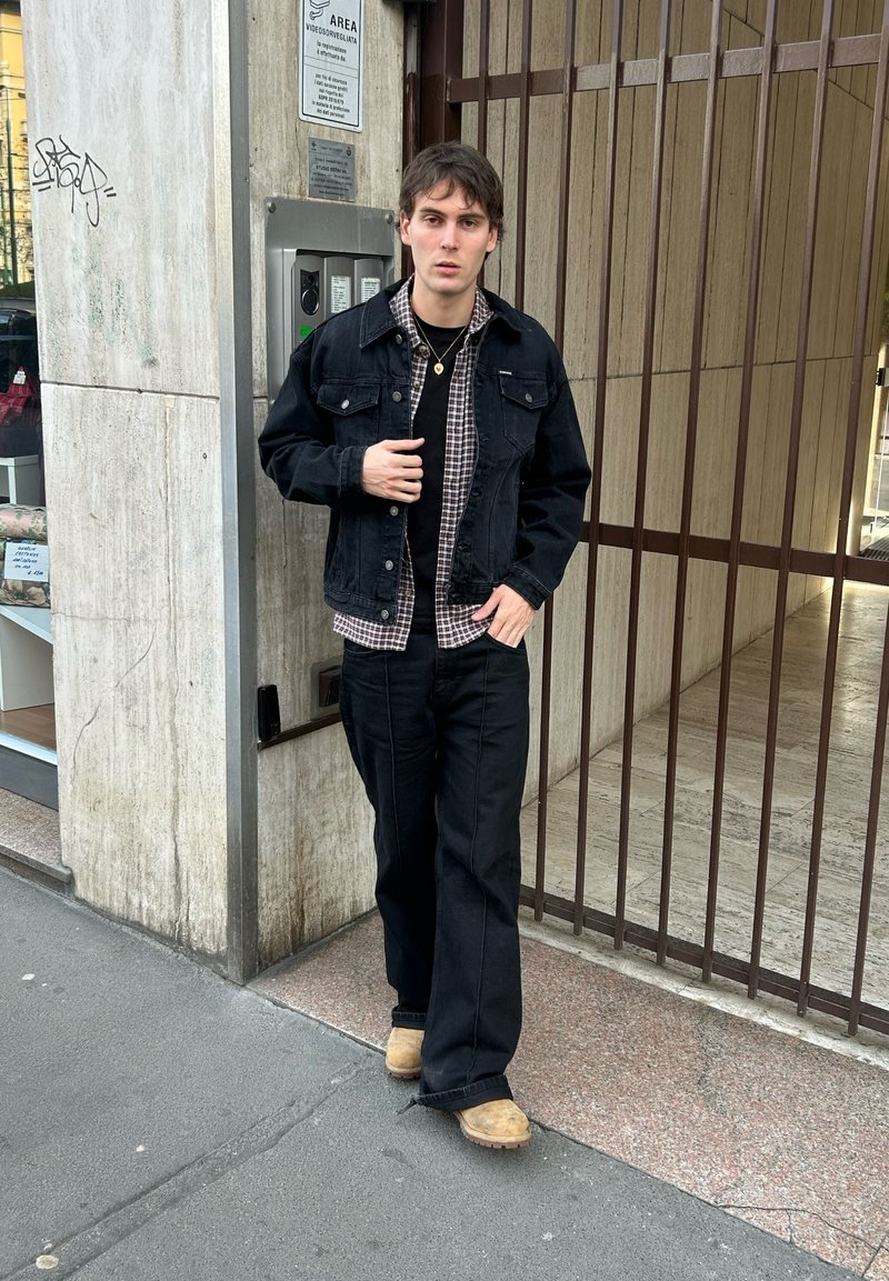 Mladý muž v čiernej bunde, kockovanej košeli, čiernych nohaviciach a hnedej obuvi stojaci pri bránovej vstupnej časti vedľa betónovej steny s nápisom.