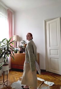 Miętowo zielony sweter z dzianiny na paskowanej koszuli, zestawiony z beżową, miękką spódnicą. W tle widać drewniane meble i rośliny doniczkowe.