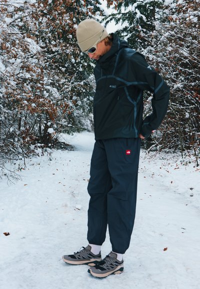 Chaqueta negra impermeable con detalles en gris, pantalones azul marino y zapatos negros y beige, en un entorno al aire libre nevado con árboles.