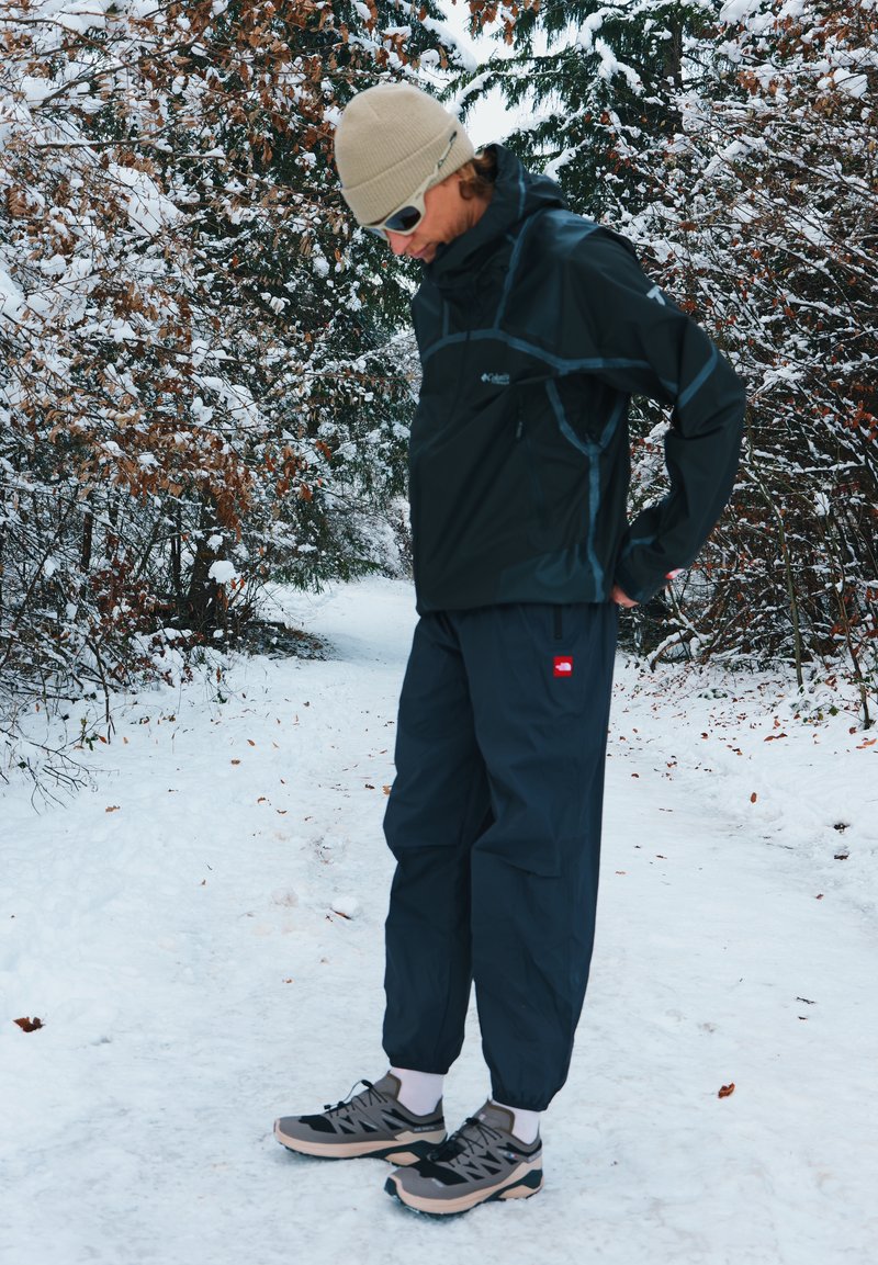 Waterdichte zwarte jas met grijze accenten, marineblauwe broek en zwart-beige schoenen, geplaatst in een besneeuwde buitenomgeving met bomen.
