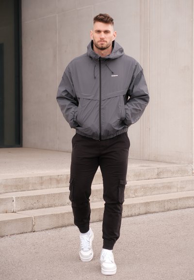 Chaqueta ligera gris con capucha, puños elásticos y cremallera frontal. Combinada con pantalones cargo negros y zapatillas blancas. Textura suave.