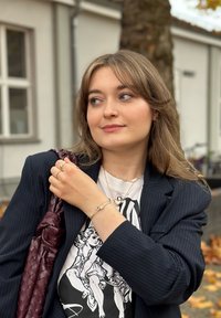 Figura femminile che indossa un blazer a righe blu navy, una t-shirt bianca con grafica e collane d'oro sovrapposte, mentre tiene una borsa a rete bordeaux.