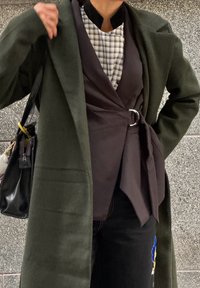 Olīvzaļš vilnas mētelis, pāri tumšam vestim un raibai blūzei, kombinēts ar tumšām džinsām ar krāsainiem ziedu izšuvumiem.