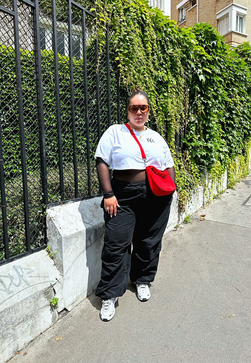 Λευκό cropped t-shirt με λογότυπο, μαύρο φαρδύ παντελόνι, κόκκινη τσάντα χιαστί και λευκά αθλητικά παπούτσια. Το μοντέλο φοράει υπερμεγέθη γυαλιά ηλίου και στέκεται δίπλα σε πράσινη βλάστηση.
