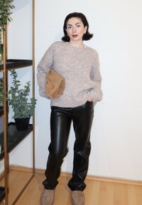 Femme aux cheveux courts et foncés portant un pull beige, un pantalon en cuir noir et des chaussures beiges, tenant une pochette marron clair, debout près d'une étagère à plantes.