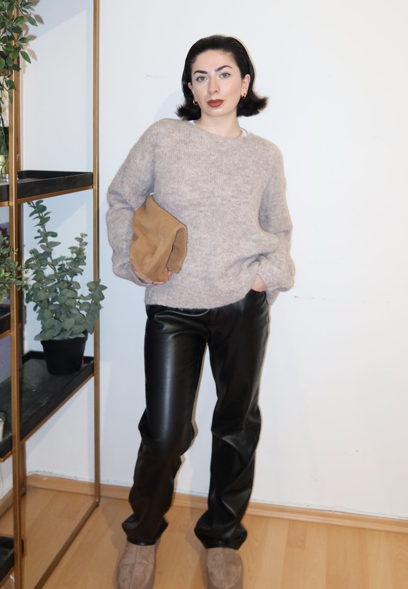 Femme aux cheveux courts et foncés portant un pull beige, un pantalon en cuir noir et des chaussures beiges, tenant une pochette marron clair, debout près d'une étagère à plantes.