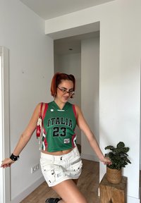 Πράσινη μπασκετική φανέλα με κόκκινες λεπτομέρειες και γράμματα "ITALIA 23", συνδυασμένη με άσπρη βερμούδα. Τα αξεσουάρ περιλαμβάνουν μια κόκκινη τσάντα και γυαλιά ηλίου.