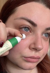 Grønn og hvit sylindrisk applikatortube holdt nær øyet, som påfører en klar gel på huden; modellen har lys hud og rødt hår.