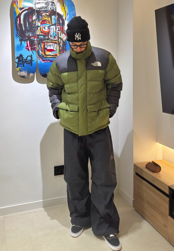 Persona che indossa una giacca verde e nera North Face, un berretto nero, pantaloni larghi neri e sneakers, in piedi al chiuso vicino a un'opera d'arte colorata con skateboard.