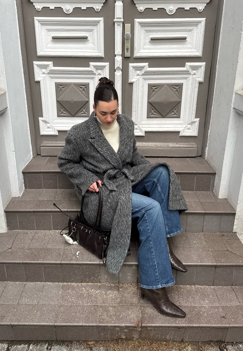 Femme en manteau gris ceinturé et jean bleu assise sur des marches marron devant des portes doubles blanches décoratives, tenant un sac à main sombre.