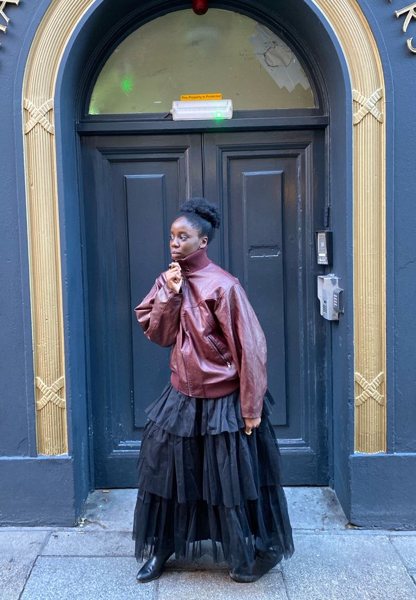 Veste en cuir marron à col montant, jupe en tulle noir superposée, et bottines noires, posée devant une porte bleu foncé avec des accents dorés.