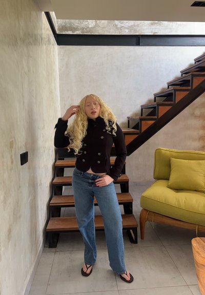 Persona con cabello largo, rubio y rizado, chaqueta negra, vaqueros azules y chanclas, de pie junto a una escalera de madera y una silla con cojines amarillos.