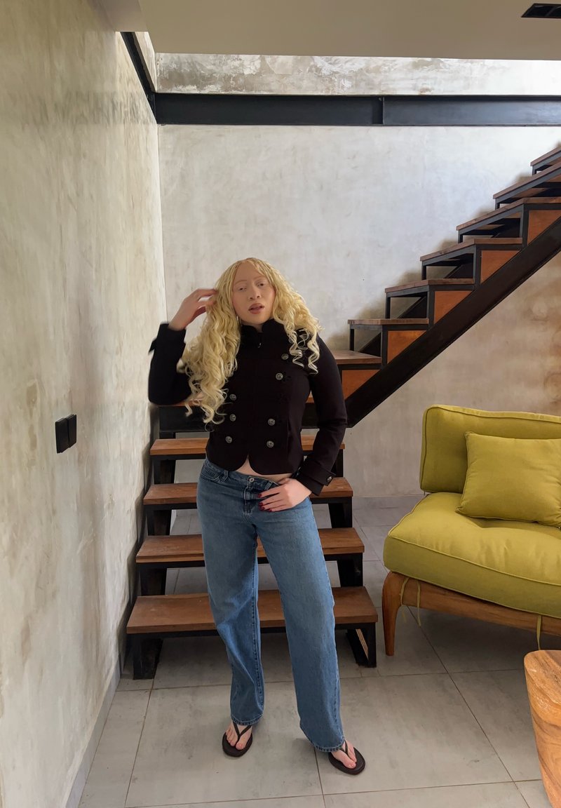 Άτομο με μακριά ξανθά σγουρά μαλλιά, μαύρο μπουφάν, μπλε τζιν και σαγιονάρες στέκεται δίπλα σε ξύλινη σκάλα και κίτρινη καρέκλα με μαξιλάρι.