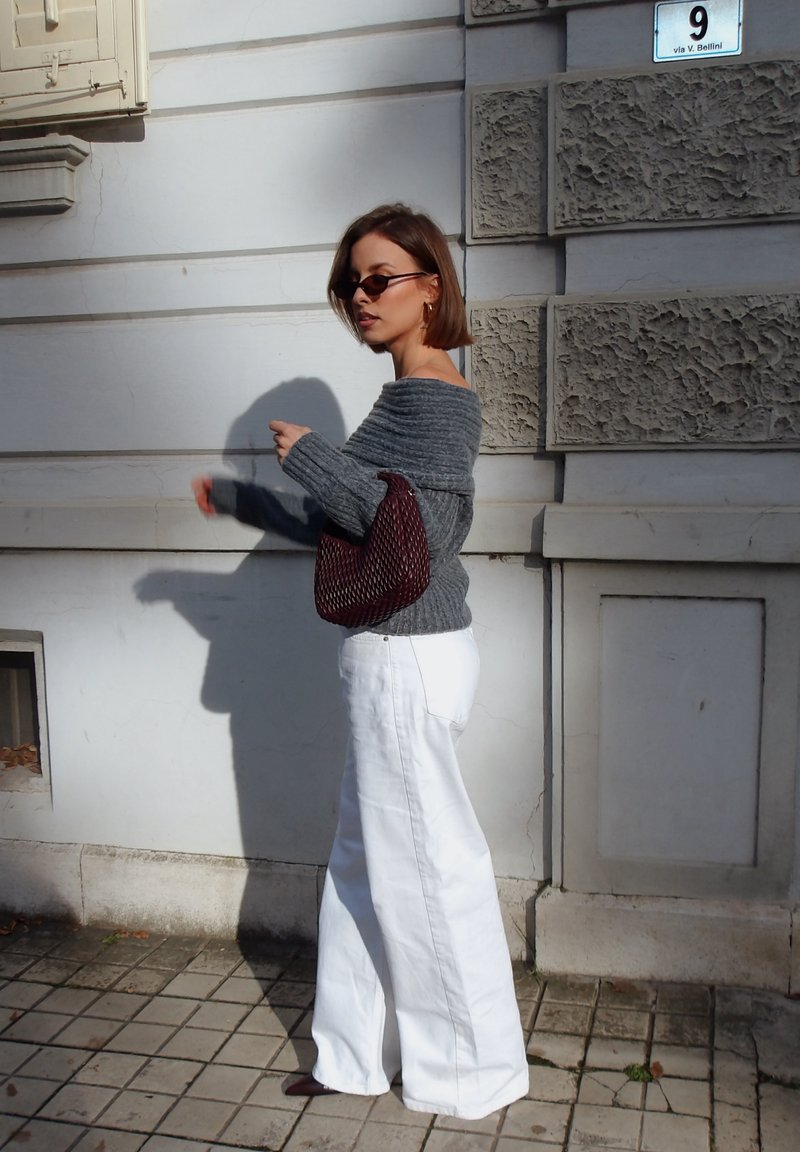 Pull gris tricoté à épaules dénudées, pantalon blanc à jambe large, et sac à main tissé bordeaux. Coupe au carré longue, lunettes de soleil rondes, et fond neutre.