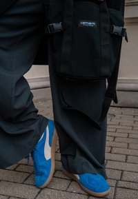 Sac en toile noire avec boucles, porté sur la hanche. Baskets en daim bleu avec des accents blancs et une semelle en caoutchouc. Pantalon gris ample. Pavé texturé en dessous.