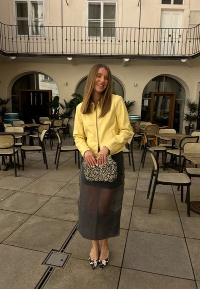 Mujer con chaqueta amarilla y falda negra transparente está de pie sonriendo en una cafetería al aire libre, sosteniendo un bolso de mano texturizado, rodeada de sillas y mesas vacías.