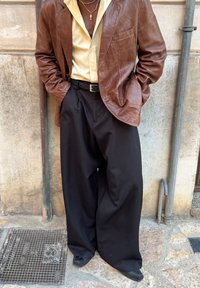 Jaqueta de couro castanha sobre uma camisa creme, combinada com calças largas pretas e um cinto preto. Acessórios com colares em camadas.