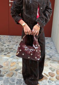 Sac à main bordeaux avec des accents d'œillets argentés, finition brillante et forme incurvée unique, associé à une tenue noire surdimensionnée posée sur un sol en pierre.