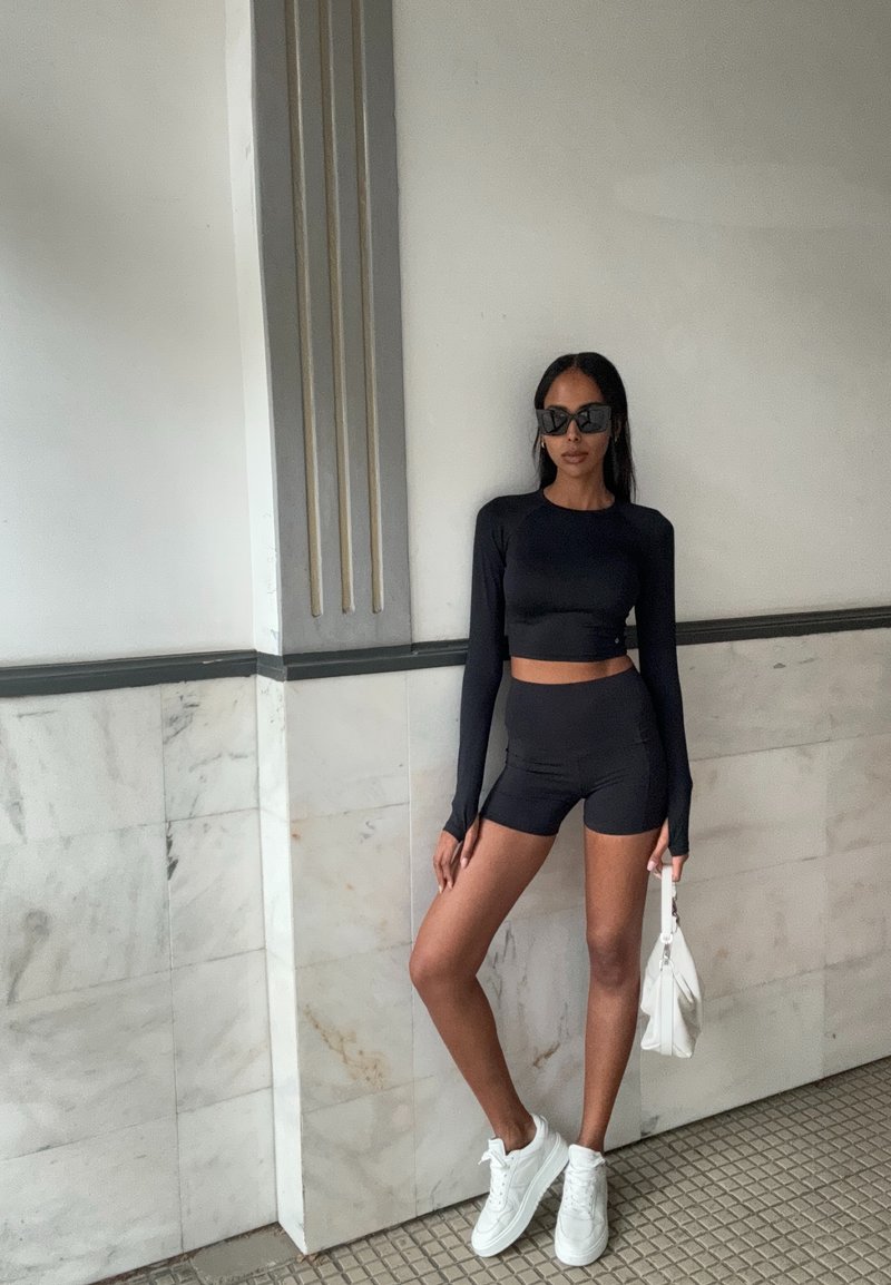 Μαύρο μακρυμάνικο crop top με στενές μαύρες βερμούδες, συνδυασμένο με λευκά αθλητικά παπούτσια και μια μικρή λευκή τσάντα. Λείο υλικό, απλός σχεδιασμός.